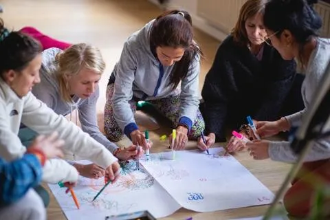 A group of women kneeling on a wooden floor together, collaboratively drawing and writing on a large sheet of white paper using colorful markers.