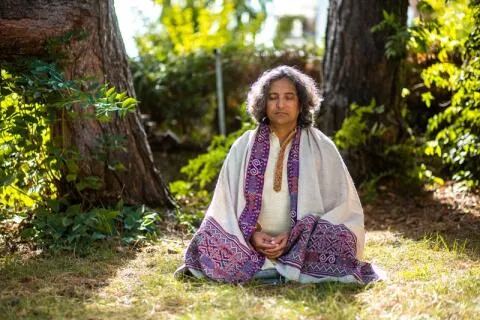 A man meditating outdoors while wearing a traditional white shawl with an embroidered purple border.
