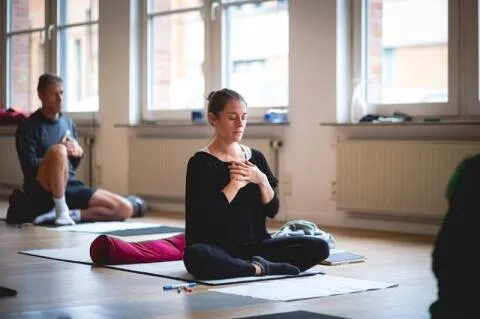 Woman sitting cross-legged in a meditation class with hands over her heart and eyes closed.