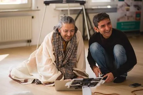 Two men smiling and sitting on a wooden floor, looking at a laptop and tablet together.