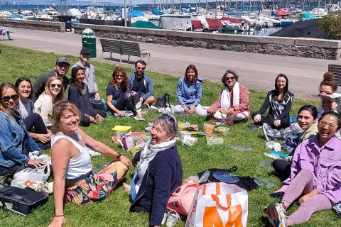 A large group of people having a picnic on a grassy lawn near a marina