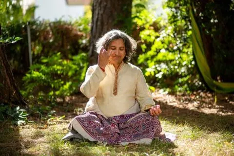  A man with shoulder-length grey hair sitting in a cross-legged meditative pose in a sunlit garden, practicing a hand mudra.