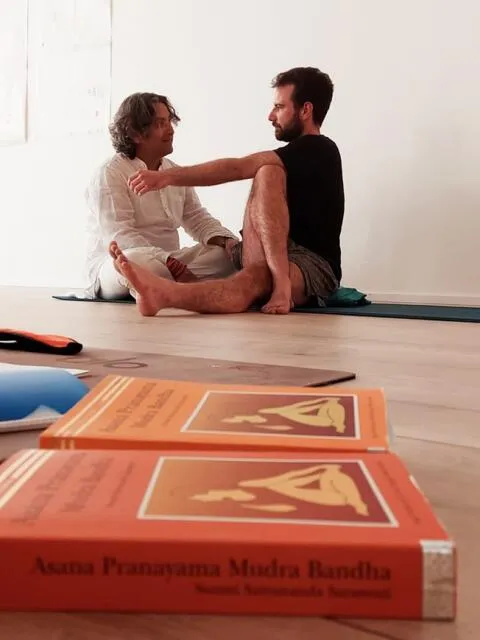 Two men in a yoga session with 'Asana Pranayama Mudra Bandha' books in the foreground.